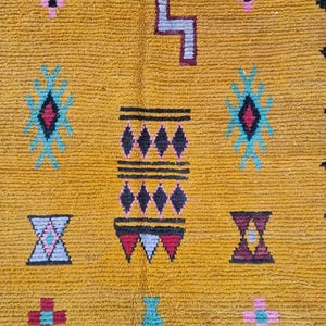 May include: Mustard yellow rug with geometric patterns in teal, pink, black, and burgundy. The rug features diamond, cross, and zigzag designs. The texture of the rug is visible, suggesting a woven or knotted construction. The rug is likely handmade.
