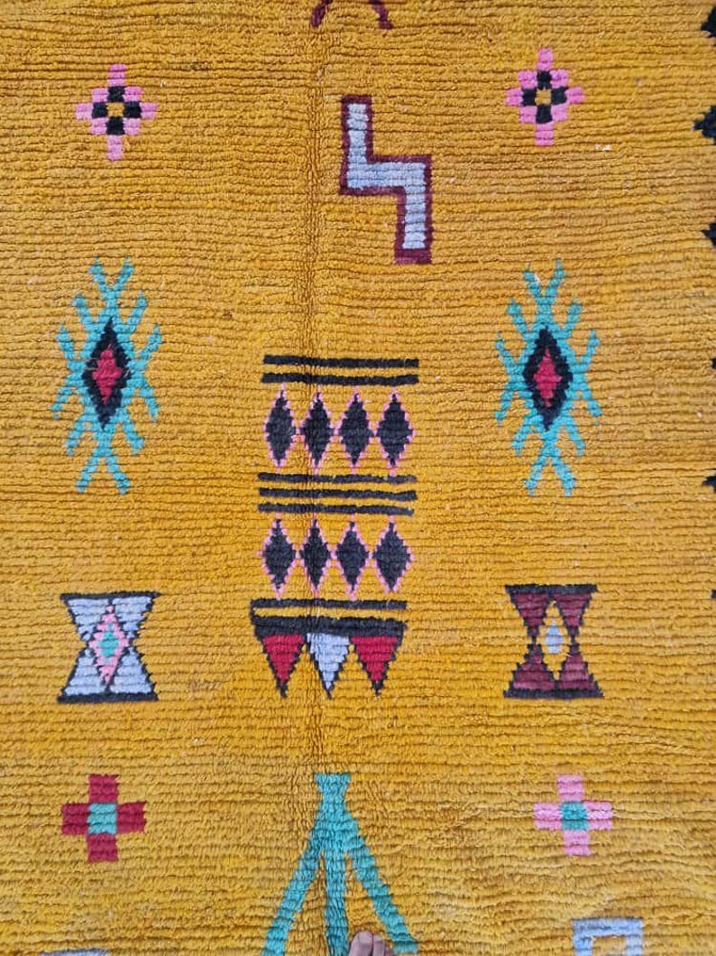 May include: Mustard yellow rug with geometric patterns in teal, pink, black, and burgundy. The rug features diamond, cross, and zigzag designs. The texture of the rug is visible, suggesting a woven or knotted construction. The rug is likely handmade.