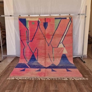 Puede incluir: Una alfombra rectangular grande con un diseño geométrico. La alfombra presenta un fondo rosa con formas azules, verdes y blancas. La alfombra tiene flecos a lo largo del borde inferior y se exhibe sobre un piso de madera.