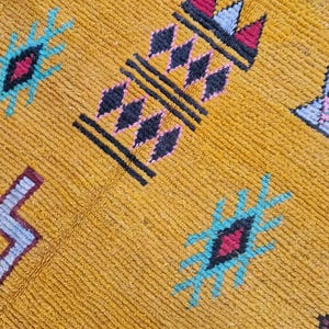 May include: Close-up of a mustard-yellow rug with geometric patterns. The design includes black, pink, red, and turquoise shapes. The rug has a textured surface and a woven appearance, suggesting a handmade or artisanal quality.