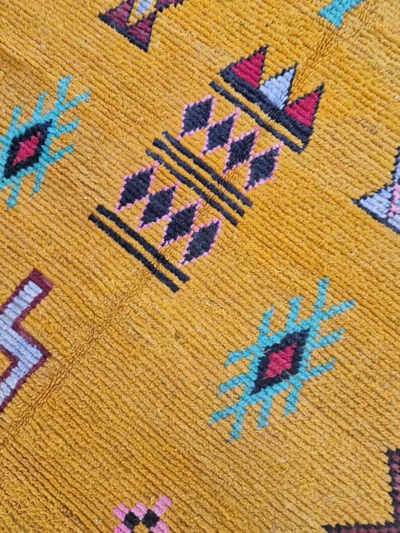 May include: Close-up of a mustard-yellow rug with geometric patterns. The design includes black, pink, red, and turquoise shapes. The rug has a textured surface and a woven appearance, suggesting a handmade or artisanal quality.