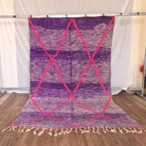 May include: A large, rectangular rug with a purple and pink geometric design. The rug features a gradient of purple hues and is accented with bright pink diamond shapes. The bottom edge has a fringe of white tassels.