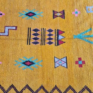 May include: Mustard yellow rug with a geometric pattern. The rug features black zigzag borders and various colorful shapes, including diamonds, crosses, and starbursts in teal, pink, and burgundy. The rug has a textured surface.