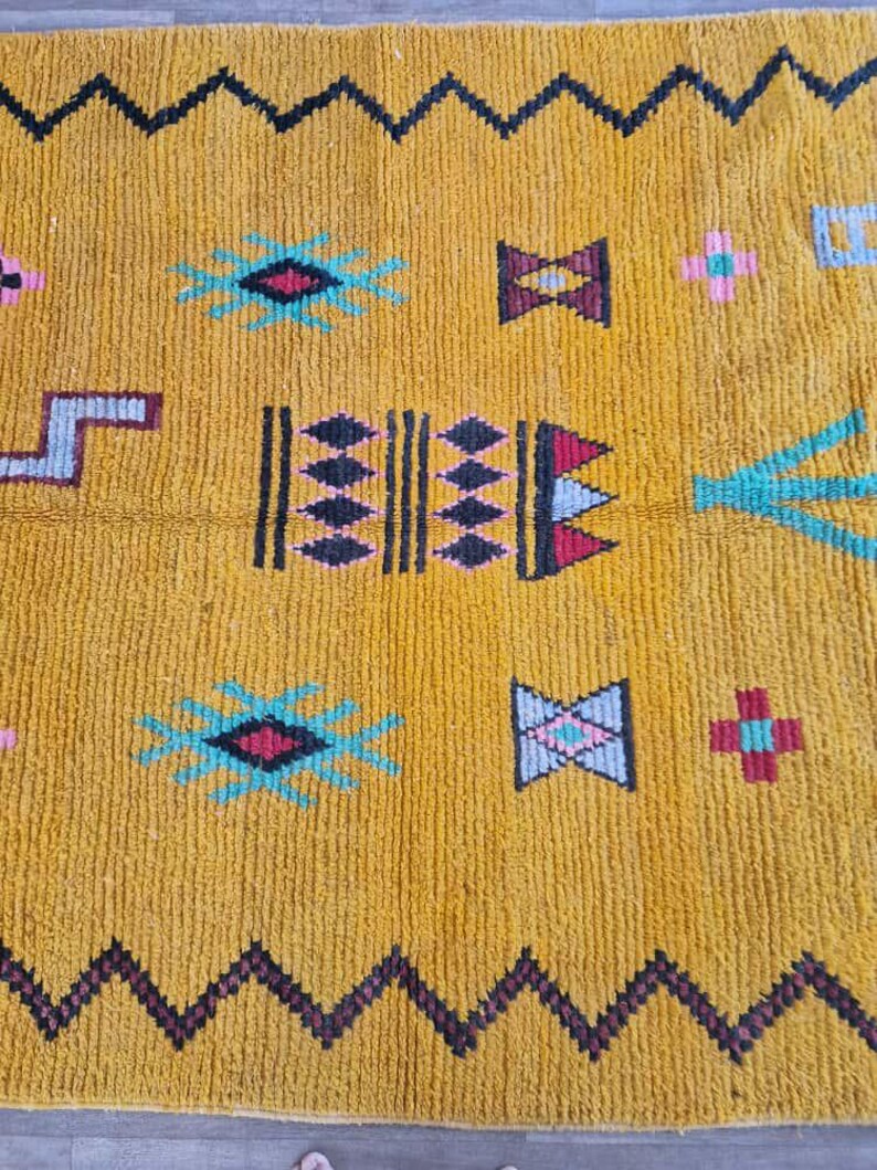 May include: Mustard yellow rug with a geometric pattern. The rug features black zigzag borders and various colorful shapes, including diamonds, crosses, and starbursts in teal, pink, and burgundy. The rug has a textured surface.