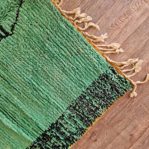 May include: A close-up of a green and black rug with a textured surface. The rug has a geometric design and fringe along the edge. The rug is made of a soft material and is suitable for home decor.