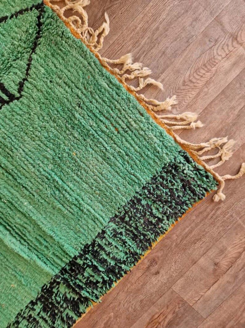 May include: A close-up of a green and black rug with a textured surface. The rug has a geometric design and fringe along the edge. The rug is made of a soft material and is suitable for home decor.