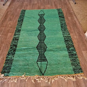 May include: A rectangular, hand-woven rug with a teal background and a central black geometric design. The rug has a black border and cream-colored fringe along the short ends. The rug is placed on a wooden floor.
