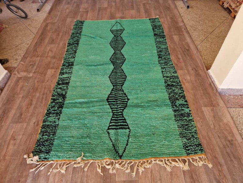 May include: A rectangular, hand-woven rug with a teal background and a central black geometric design. The rug has a black border and cream-colored fringe along the short ends. The rug is placed on a wooden floor.