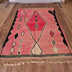 May include: A rectangular pink rug with a geometric design. The rug features a large diamond shape in the center, with smaller diamond and other shapes in black, white, and green. The rug has a fringe along the bottom edge.
