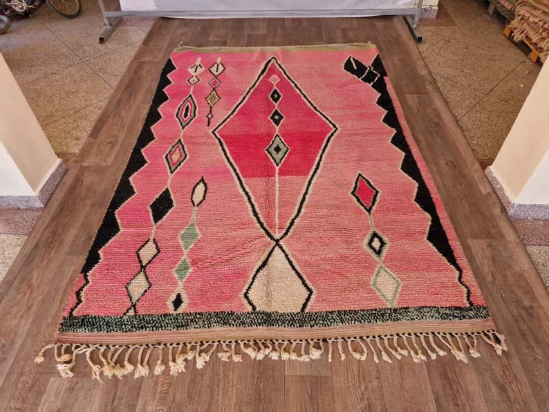 May include: A rectangular pink rug with a geometric design. The rug features a large diamond shape in the center, with smaller diamond and other shapes in black, white, and green. The rug has a fringe along the bottom edge.
