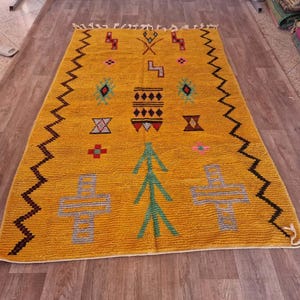 May include: Mustard yellow rug with geometric patterns in various colors. The rug features a zig-zag border in dark brown and black, with symbols in green, red, and blue. The rug is rectangular and appears to be made of a textured material.