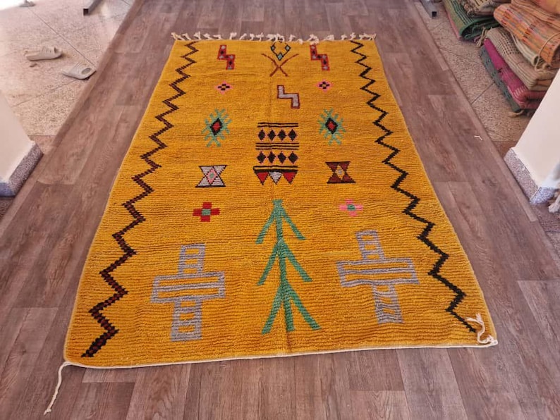 May include: Mustard yellow rug with geometric patterns in various colors. The rug features a zig-zag border in dark brown and black, with symbols in green, red, and blue. The rug is rectangular and appears to be made of a textured material.