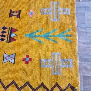 May include: Mustard yellow rug with geometric patterns. The rug features black zigzag borders, teal and gray cross shapes, and various other colorful geometric designs. The rug has a textured appearance and a neutral background.