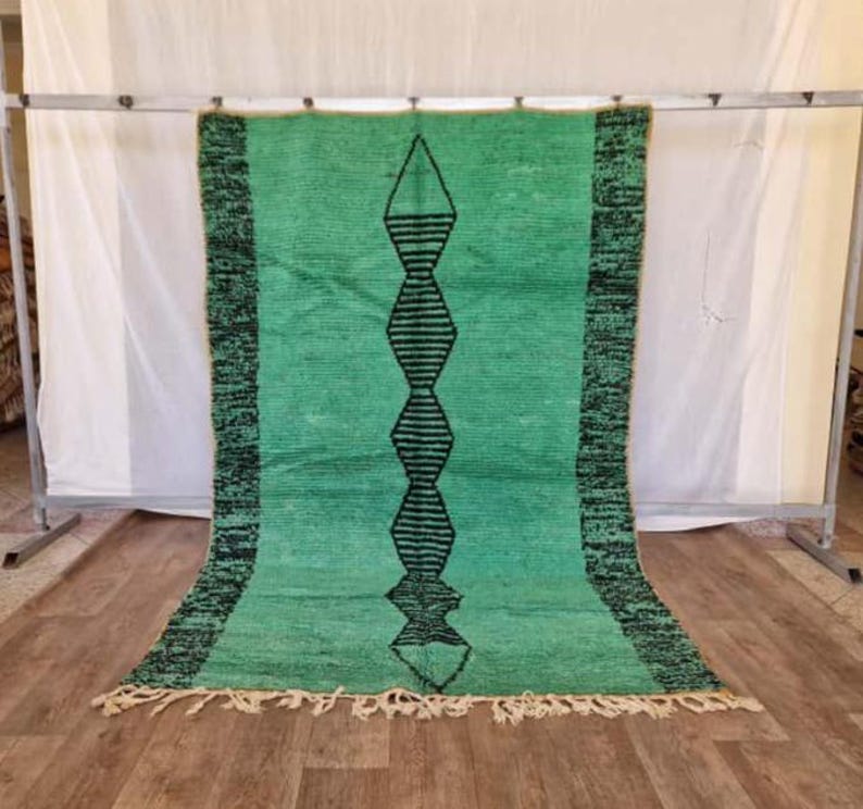May include: A rectangular, hand-woven rug in shades of green with a central black geometric design. The rug has a fringe along the bottom edge and a dark green border. The rug is displayed against a white backdrop.