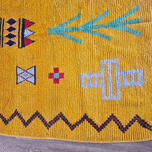May include: Close-up of a mustard yellow rug with geometric patterns. The rug features black, red, and blue diamond shapes, a zig-zag border, and teal and white arrow and cross designs.
