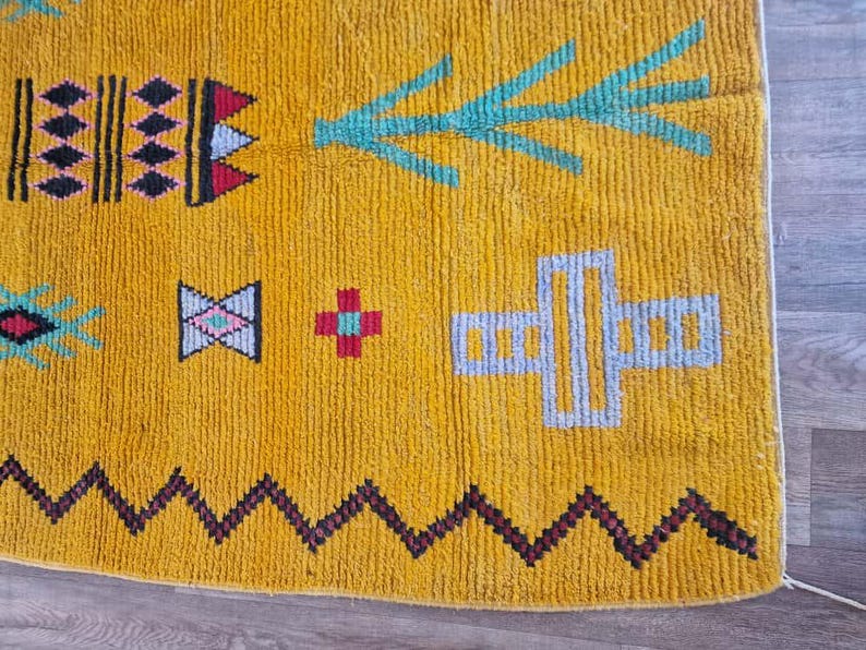 May include: Close-up of a mustard yellow rug with geometric patterns. The rug features black, red, and blue diamond shapes, a zig-zag border, and teal and white arrow and cross designs.