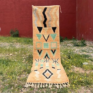 Peut inclure: Tapis rectangulaire tissé à la main, fond beige et motifs géométriques noirs, turquoise et orange. Le tapis présente un motif en zigzag, des triangles, des carrés et des losanges. Il a une frange en bas et est exposé à l'extérieur.