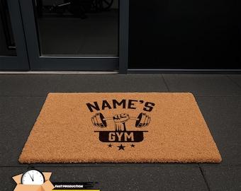 May include: A rectangular brown coir doormat with the text "NAME'S GYM" above a graphic of a weightlifter's hand holding a barbell. The word "GYM" is below the graphic, with three stars beneath. The mat is on a black floor.