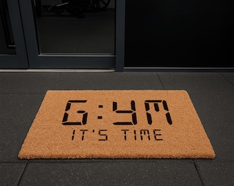 May include: A rectangular doormat made of natural brown coir fiber. The mat features the word "GYM" in a digital clock font, with the phrase "IT'S TIME" below it in black lettering. The mat is placed on a black floor.