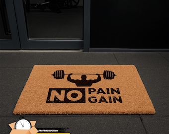 May include: A rectangular doormat made of natural brown coir fiber. The mat features a black graphic of a weightlifter lifting a barbell, with the words "NO PAIN GAIN" in black block letters. The mat is placed on a black floor.