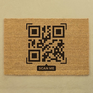 May include: A rectangular doormat made of natural brown coir fibre. A large black QR code is printed in the centre, with the words "SCAN ME" in a white rectangle below the code. The mat is on a light beige surface.