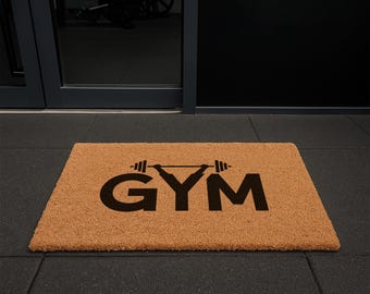 May include: A rectangular doormat with the word "GYM" in large, black capital letters. Above the "Y" is a barbell graphic. The mat is a natural tan color, made of a coarse material, and sits on a dark gray floor.
