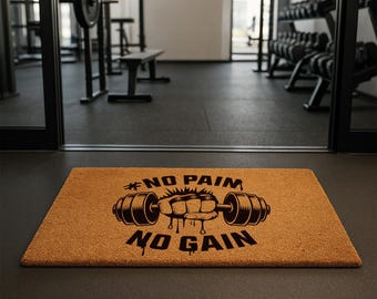 May include: A rectangular doormat with a brown surface and black text and graphics. The text reads "#NO PAIN NO GAIN" with a graphic of a weightlifting barbell and a clenched fist. The doormat is in a gym setting.