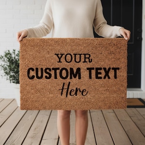 Puede incluir: Una felpudo rectangular de fibra de coco marrón con el texto "YOUR CUSTOM TEXT Here" en negro. La alfombra es sostenida por una persona frente a una pared de ladrillos blancos y una puerta negra.