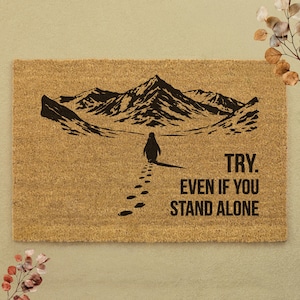 Könnte beinhalten: Eine rechteckige Fußmatte mit einem Pinguin, der auf eine Bergkette zugeht. Die Matte ist braun mit einer schwarzen Illustration und dem Text "TRY. EVEN IF YOU STAND ALONE."