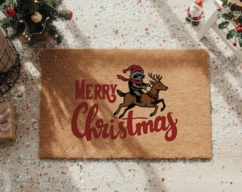 May include: A rectangular doormat with a brown coir surface. The mat features the words "Merry Christmas" in red script, along with an illustration of a raccoon riding a reindeer. The raccoon wears a red hat and scarf.