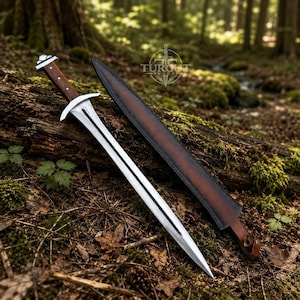 Puede incluir: Una espada de doble filo con hoja de acero pulido, guarda plateada y empuñadura de madera marrón oscuro con remaches metálicos. Junto a la espada hay una funda de cuero marrón oscuro a juego con el texto TUROUT BLADES.