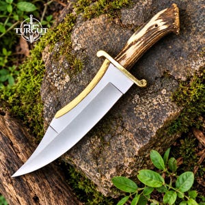 Puede incluir: Un cuchillo de hoja fija con una hoja de acero plateado pulido, una guarda de metal dorado curvada y un mango de asta de ciervo marrón texturizado. La hoja presenta una punta afilada y un filo curvo. El texto TURGUT BLADES es visible en la parte superior izquierda.