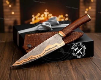 May include: A Damascus steel chef's knife with a wooden handle and a leather sheath. The blade has a wavy pattern with gold accents. The knife is presented in a black gift box with a white ribbon.