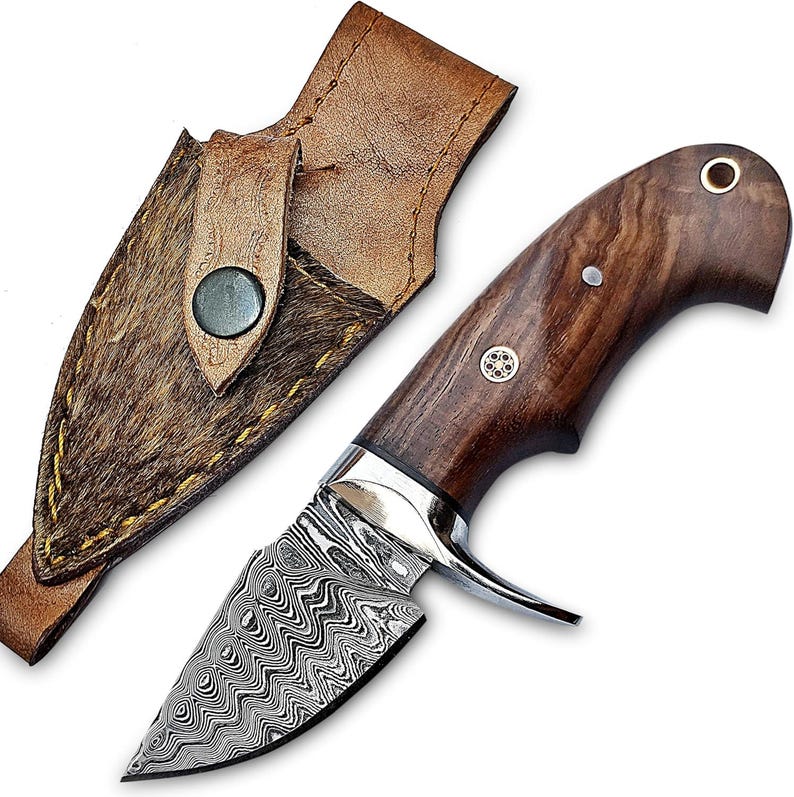 May include: A fixed-blade knife with a patterned Damascus steel blade and a dark brown wood handle featuring a decorative metal pin. The knife is accompanied by a brown leather sheath with a textured hair-on-hide inlay and a metal snap closure.