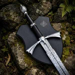 May include: A sword with a patterned Damascus steel blade, a black leather-wrapped handle, and a silver-toned pommel and crossguard. The sword is mounted on a black shield-shaped plaque featuring the word TURGUT printed in white.