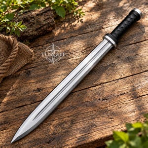 May include: A double-edged steel sword with a central fuller and a black wrapped handle. The blade features a silver finish and a pointed tip. A logo with the text TURGUT BLADES is visible on the blade surface near the hilt of the weapon.