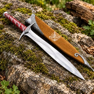 May include: A silver-toned metal dagger with a red handle featuring silver leaf-like patterns and a matching crossguard. The blade has intricate engravings. Beside it lies a brown suede scabbard with silver metal accents and the text TURGUT BLADES.