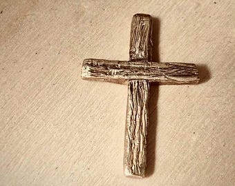 May include: A silver-colored cross with a textured, wood-like appearance. The cross is a simple design, with a vertical and horizontal beam intersecting at right angles. The surface has a rough, aged look.