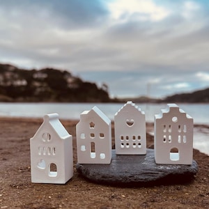 Puede incluir: Cuatro pequeñas figuras de casas de cerámica blanca con varios recortes de ventanas y puertas están dispuestas sobre una piedra gris oscura. Las casas se encuentran sobre un fondo borroso de una playa, agua y un cielo nublado.