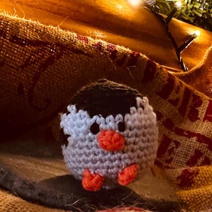 Puede incluir: Una figura de pingüino hecha a mano con ganchillo. El pingüino tiene la cabeza negra, el cuerpo blanco y gris, y las patas y el pico naranjas. El pingüino está sobre una superficie de arpillera, con un fondo de madera y luces de cadena.