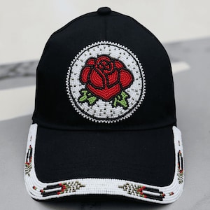 Pode incluir: Boné de beisebol preto com um design de rosa bordada. A rosa é vermelha com folhas verdes, rodeada por um círculo de contas brancas. A aba e as laterais do boné apresentam contas brancas com padrões decorativos.