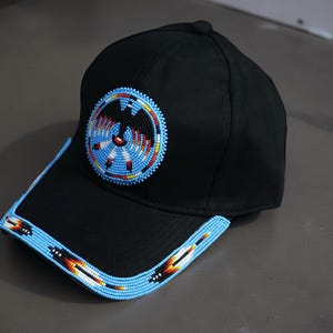 Könnte beinhalten: Schwarze Baseballkappe mit Perlendesign. Die Kappe hat einen kreisförmigen Perlenaufnäher mit blauen, roten und gelben Akzenten. Der Rand ist mit einer hellblauen Perlenborte mit geometrischen Mustern verziert.