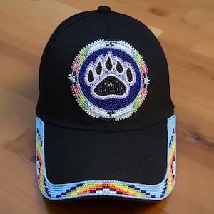 May include: Black baseball cap featuring a beaded paw print design. The paw print is within a blue circle, surrounded by white and colourful beads. The cap's brim is also adorned with colourful beads. A fashionable accessory.