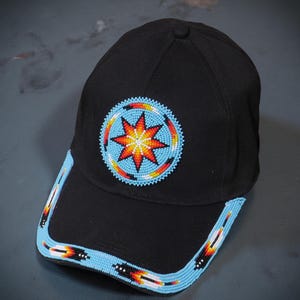Puede incluir: Gorra de béisbol negra con un diseño de cuentas. La gorra presenta un emblema circular central con un patrón de estrella en rojo, naranja y amarillo, rodeado por un borde de cuentas azules. La visera tiene un ribete de cuentas azules con patrones geométricos coloridos.