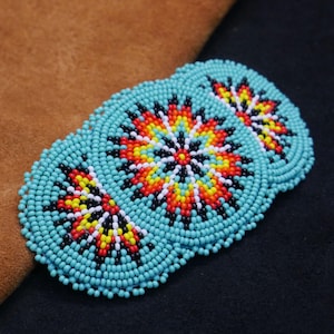 May include: Three turquoise beaded coasters with a central floral design in red, orange, yellow, and black. The coasters are round and have a detailed beaded pattern. The background is a mix of brown and black.