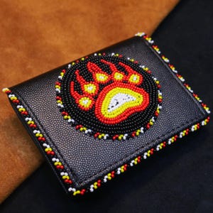 May include: Black leather wallet with a beaded bear paw design. The bear paw is in a circle, with red, yellow, and white beads. The edges of the wallet are also decorated with beads in red, yellow, and white.
