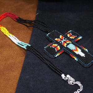 May include: A beaded cross necklace with a black background. The cross is black with a colorful design in the center, including red, orange, yellow, and turquoise beads. The necklace chain is black with a silver clasp.