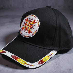 Könnte beinhalten: Schwarze Baseballkappe mit Perlendesign. Die Kappe hat ein kreisförmiges Emblem mit einem roten, gelben und weißen Blumenmuster. Der Rand ist ebenfalls mit einem Perlendesign in Rot, Gelb und Weiß verziert.