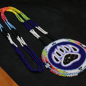 Pode incluir: Um colar de contas com um pingente circular com uma pegada de urso. O pingente é principalmente azul com uma pegada branca e um centro preto. As mechas do colar são multicoloridas, incluindo vermelho, laranja, amarelo, verde, azul e branco.
