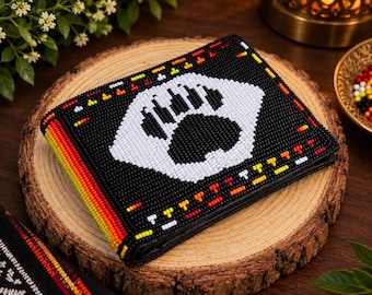 May include: A bi-fold wallet featuring intricate black, white, red, and yellow beadwork. The front displays a prominent white bear paw print design on a black background, accented by a vibrant multi-colored gradient stripe along the side.